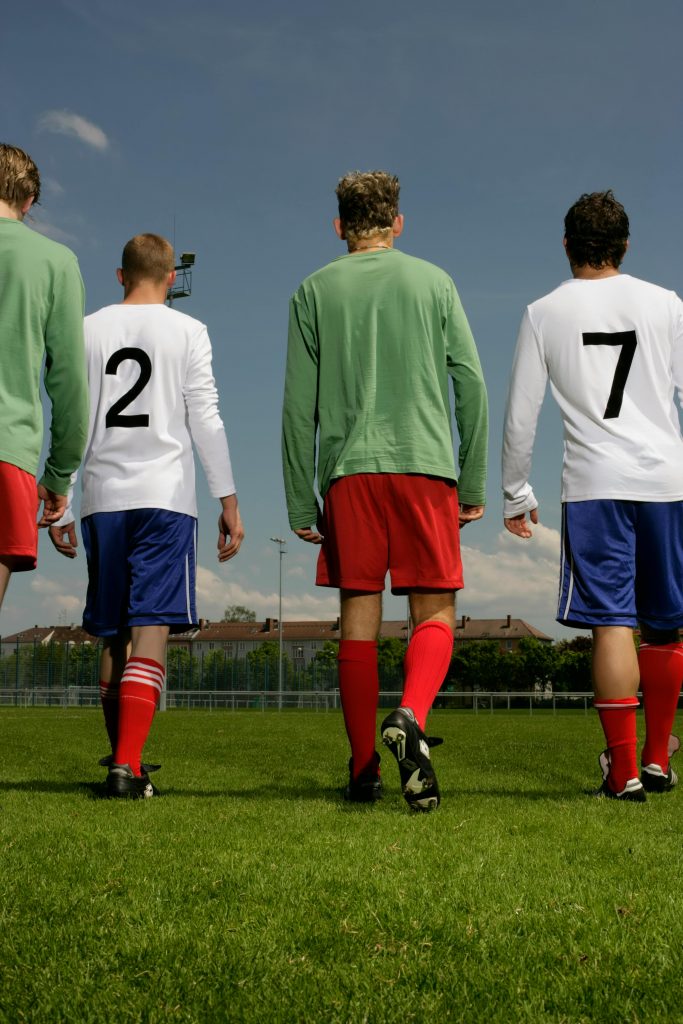 Four soccer players walking on a grassy field in bright uniforms.